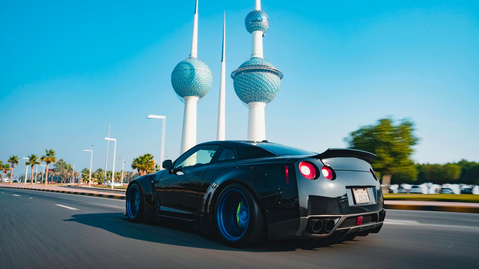 A sleek black sports car driving past the iconic Kuwait Towers on a sunny day.