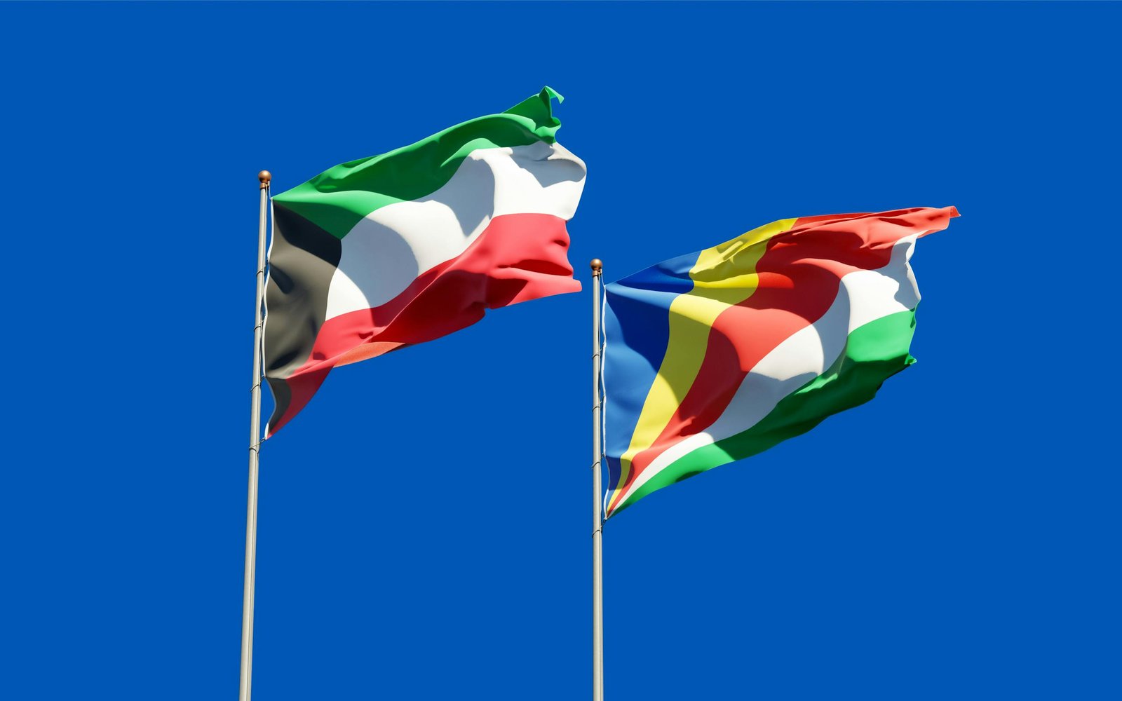 Kuwait and Seychelles national flags waving on flagpoles against a clear blue sky, symbolizing international relations.