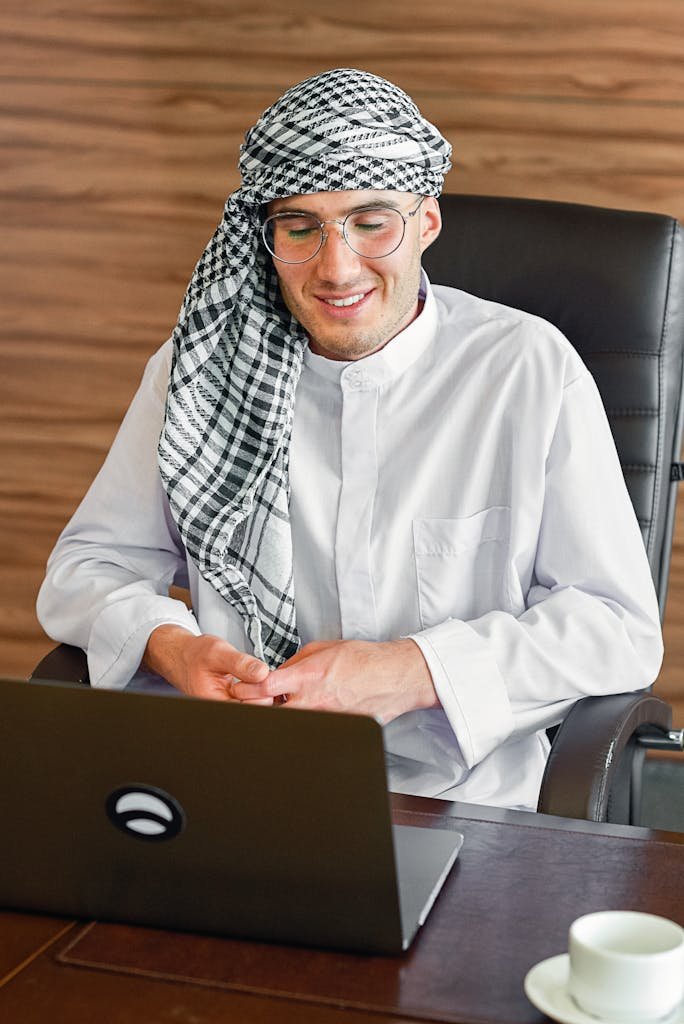 Smiling Middle Eastern man in traditional attire working on a laptop indoors.